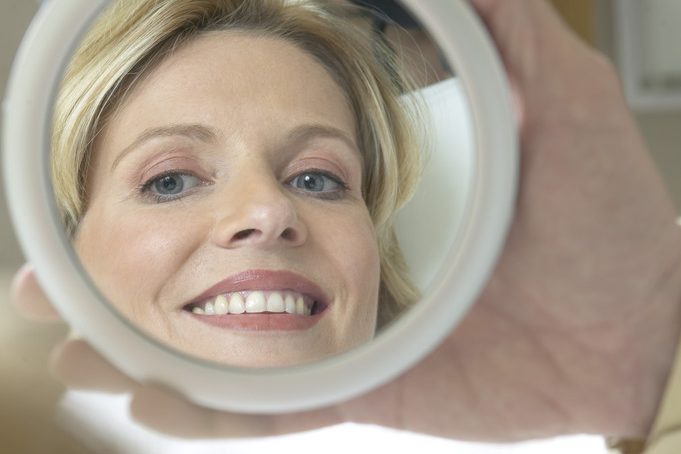 Una mano sostiene un espejo circular donde se refleja el rostro de una mujer rubia sonriente