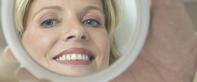 Una mano sostiene un espejo circular donde se refleja el rostro de una mujer rubia sonriente